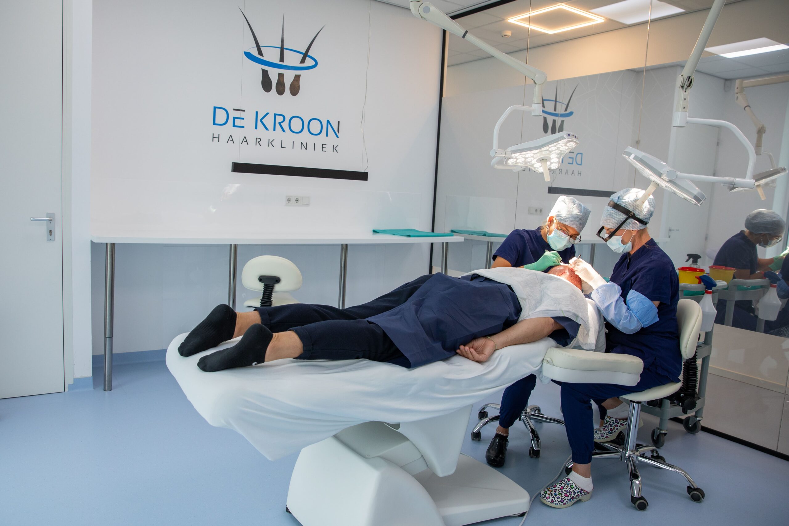 Twee specialisten voeren een haartransplantatie uit bij een patiënt die ligt op een behandelstoel in een moderne behandelkamer van De Kroon Haarkliniek.