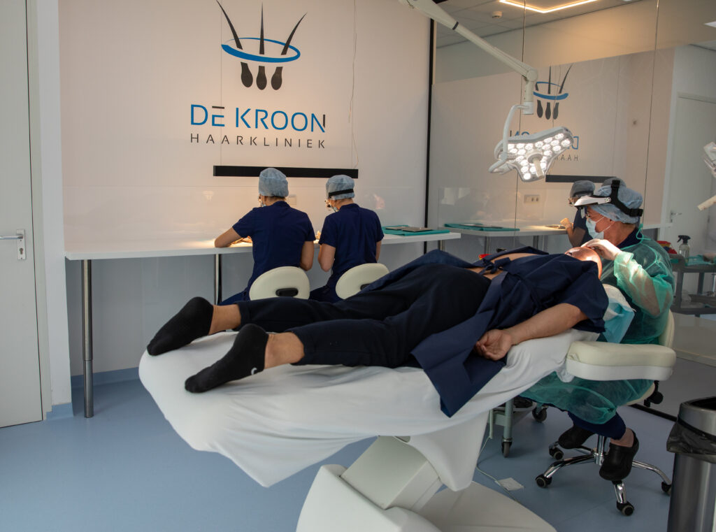 De operatiekamer tijdens een haartransplantatie bij Haarkliniek De Kroon, waarbij de cliënt ligt en de arts bezig is met de transplantatie, terwijl op de achtergrond 2 haartransplantatie specialisten bezig zijn met het verwerken van de grafts.