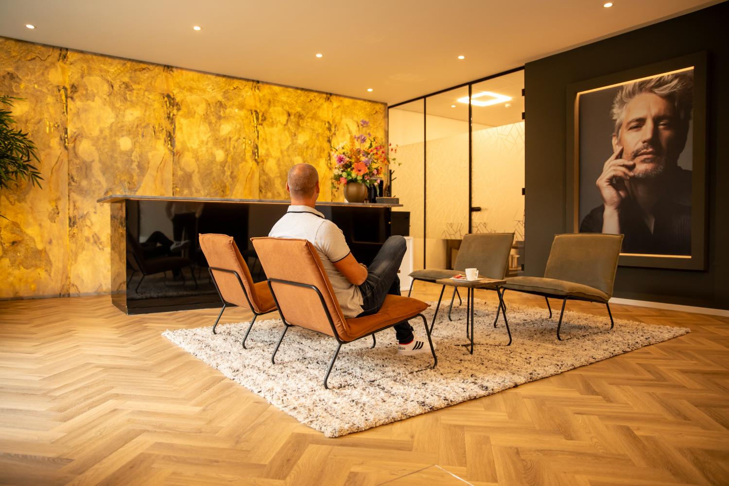 Een man zit in een moderne ontvangstruimte met designmeubilair, een gouden marmerlook wand, een zwarte balie en een groot portret aan de muur.